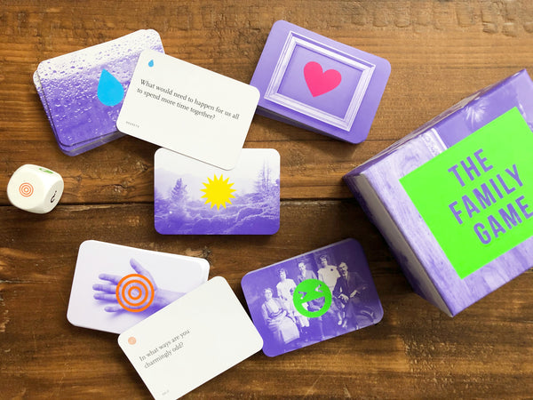 The Family Game purple and neon green product packaging lays to the side of stacked game cards and dice on a wooden background
