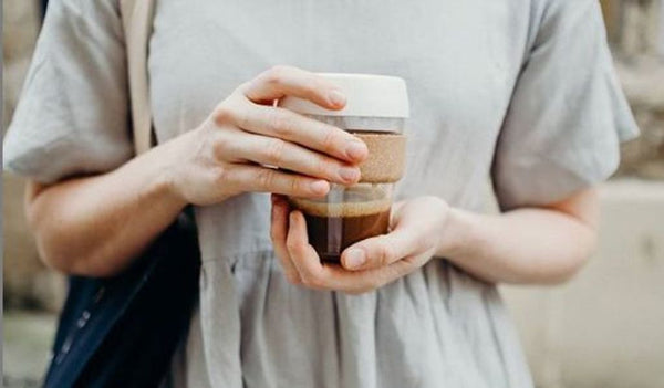 Person wearing a gray linen dress with a black tote back holding a clear glass KeepCup travel mug