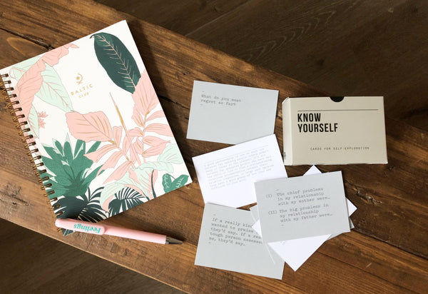 Know Yourself prompt cards lay layered on a wooden bench next to a tropical-themed illustrated notebook and pastel pink pen reading "Feelings" on the pen body