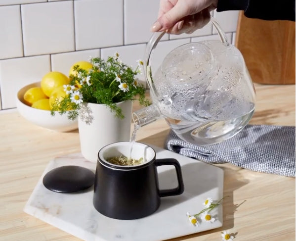 Hand pouring boiling water from a glass kettle to brew a cup of chamomile tea