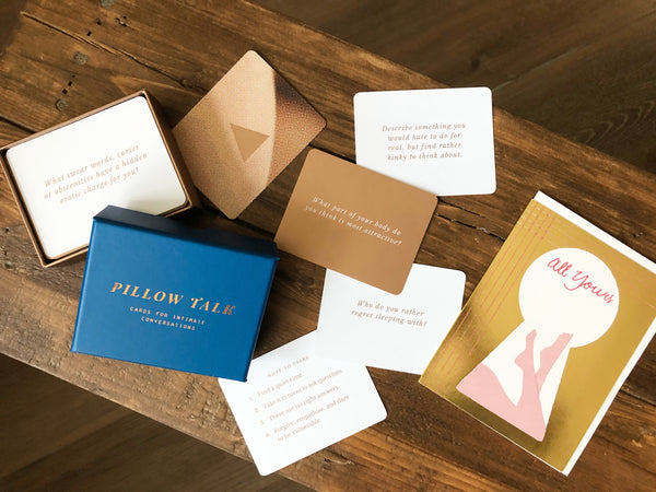 Pillow Talk conversation cards lay on a wooden bench with their royal blue storage box laying opened and a metallic greeting card reading all yours with the outline of keyhole and illustrated legs through the keyhole