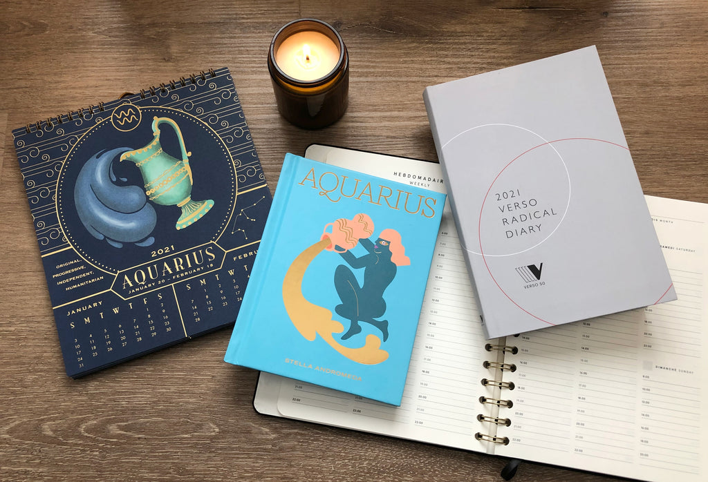A zodiac calendar displaying the months January and February lays open next to an Aquarius horoscope book and Verso Radical daily planner on a wood background