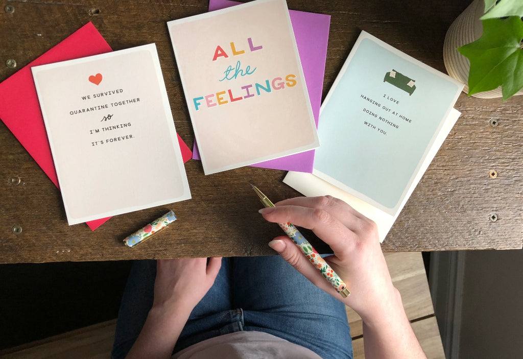 Selection of three colourful greeting cards and envelopes sit on a desk with a hand holding a floral pen to the side and a small ivy leaf peeking in the edge of the frame
