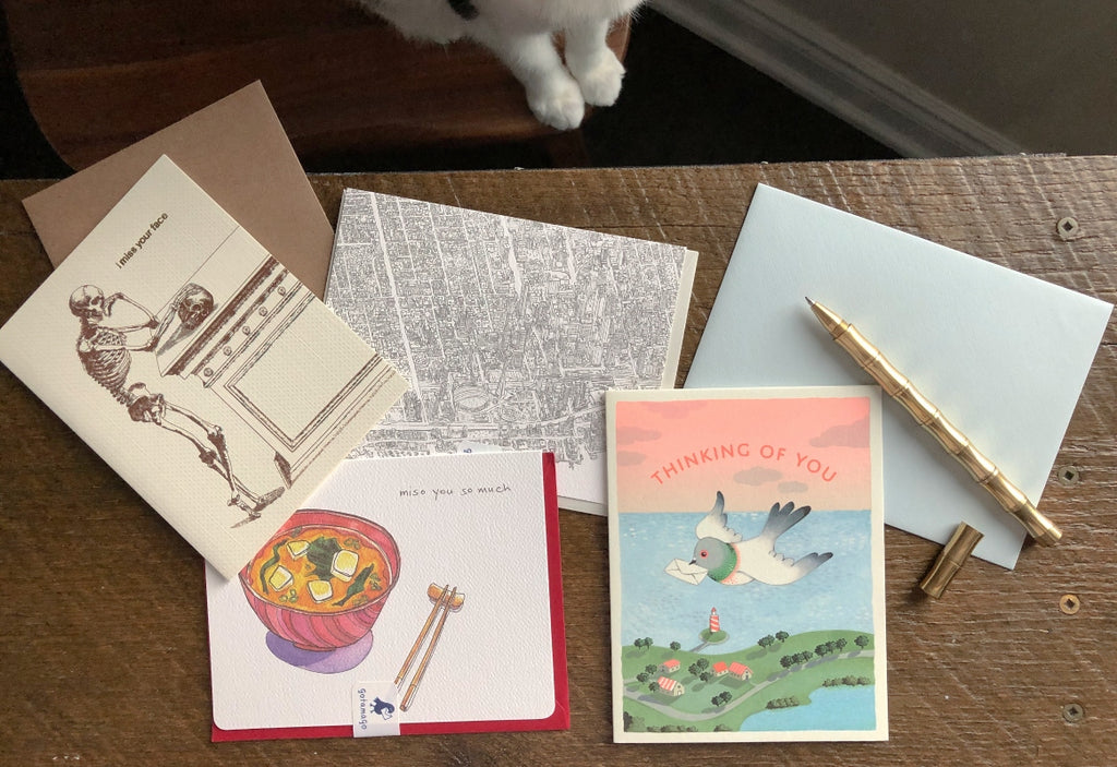 Greeting cards and envelopes lay strewn on a small wooden table with a golden pen set to the side and the white paws of a cat in the corner of the photo  