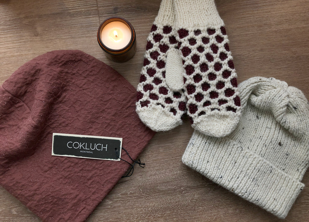 Rust-coloured slouchy toque, knit white and burgundy mittens and a rib-stitched knit ecru toque lay on a wooden background