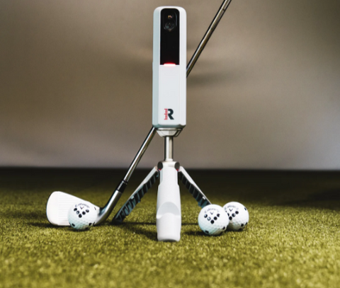 A Rapsodo MLM2 Pro launch monitor is positioned in front of a simulator, surrounded by golf clubs and balls. The simulator setup includes a screen displaying virtual golf course scenery. The Rapsodo MLM2 Pro is prominently featured, capturing swing data and providing feedback for an immersive golfing experience