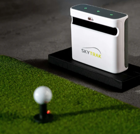 The Skytrak+ Launch Monitor is positioned on a hitting mat, featuring a ball placed on a rubber tee in front of it. The setup suggests a golf practice scenario, with the Skytrak+ ready to capture swing data. The hitting mat and rubber tee indicate a space designed for golf training and performance analysis, offering a visual representation of the equipment in use.