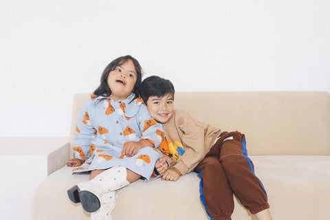 a photo of two siblings sitting on a couch. The sister has Down syndrome and is sitting on the left and the brother is leaning on her shoulder sitting on the right
