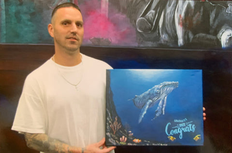 A picture of Toby holding a painting. He has close shaved hair, a 5 o'clock shadow, and tattoos on his arms. He's wearing a white shirt and is holding a painting of a while with the text "graduate 2022 congrats!".