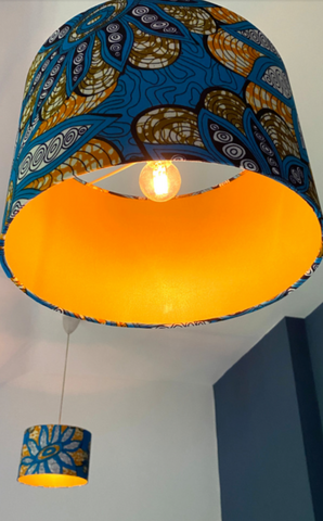 Two orange & turquoise retro flower power patterned lampshades. Both are hanging from the ceiling against a blue painted feature wall. One lampshade is in front whilst the other is in the background. You can see the inner gold lining and the light is on creating a warm glow.