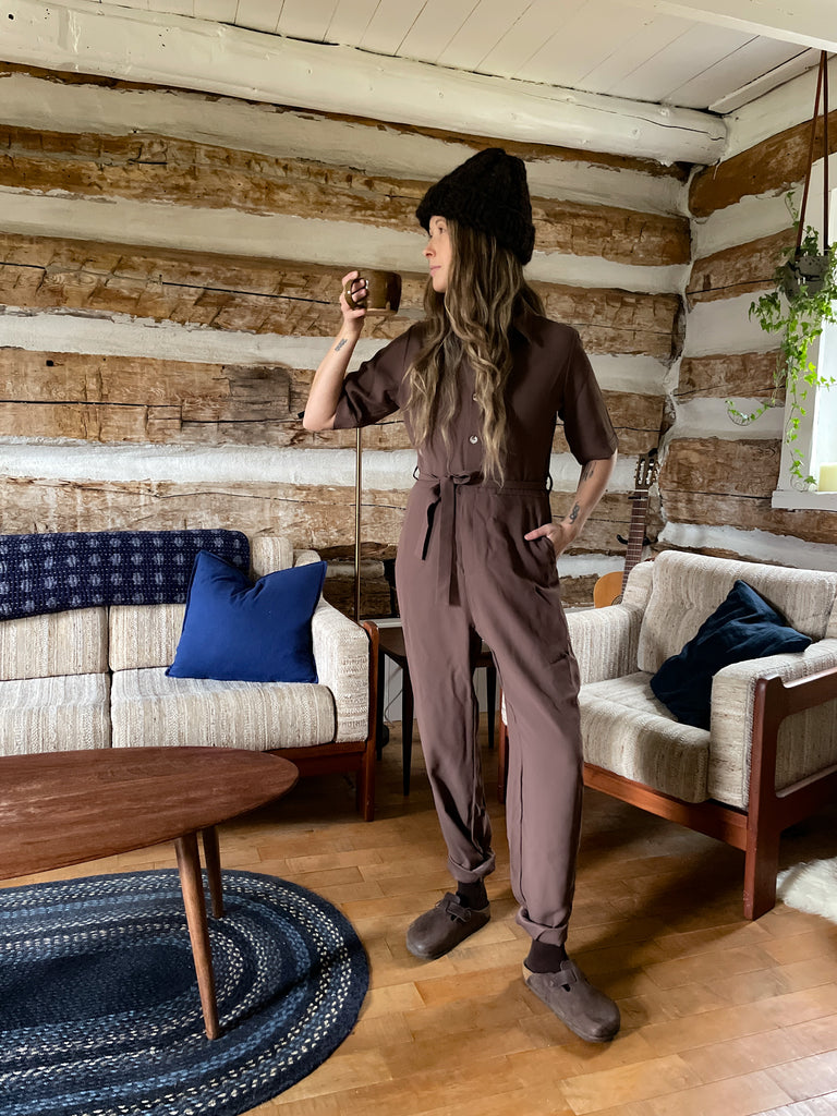 Leah, a florist, is standing up, drink our of a mug, inside a cotage living room, window in the back, looking away from the picture. A plant hangs in the back. She's wearing a The Florist Jumpsuit, Hot Chocolate colour.