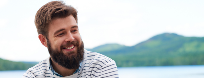 Bearded, well-groomed man smiling in front of a mountainscape wearing a striped shirt