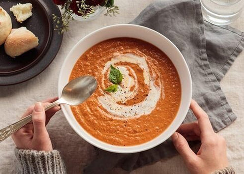 Hands holding a bowl of Spoonful of Comfort Tomato Basil Soup with rolls.