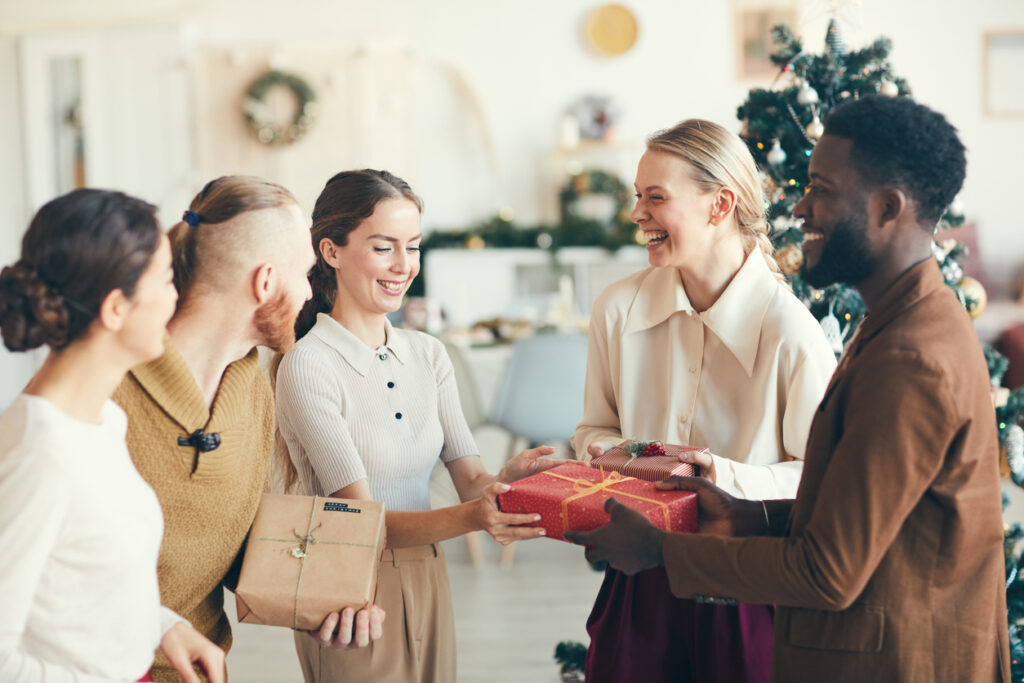 Waist up portrait of cheerful young people exchanging presents during Christmas party in cozy interior, copy space