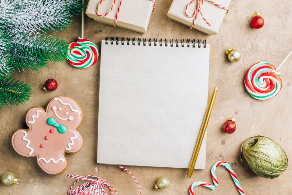 Notebook in the center of cute Christmas decoration: gift box, christmas balls, gingerbread man, fir tree branch, cande cane. Flat lay. Top view