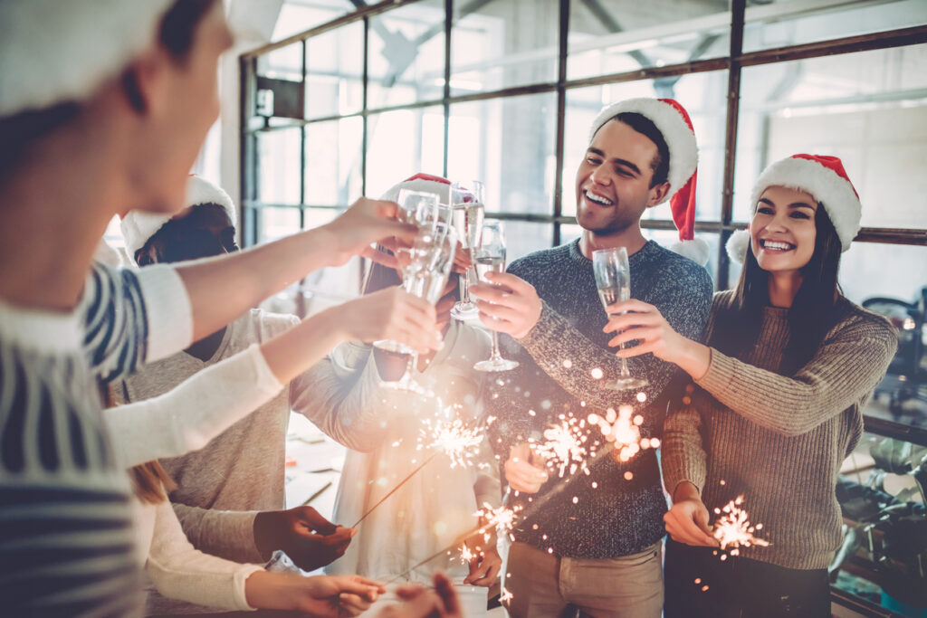 Merry Christmas and Happy New Year 2020!Multiracial young creative people are celebrating holiday in modern office. Group of young business people are drinking champagne with sparkling bengal lights in coworking. Successful hipster team of freelancers.