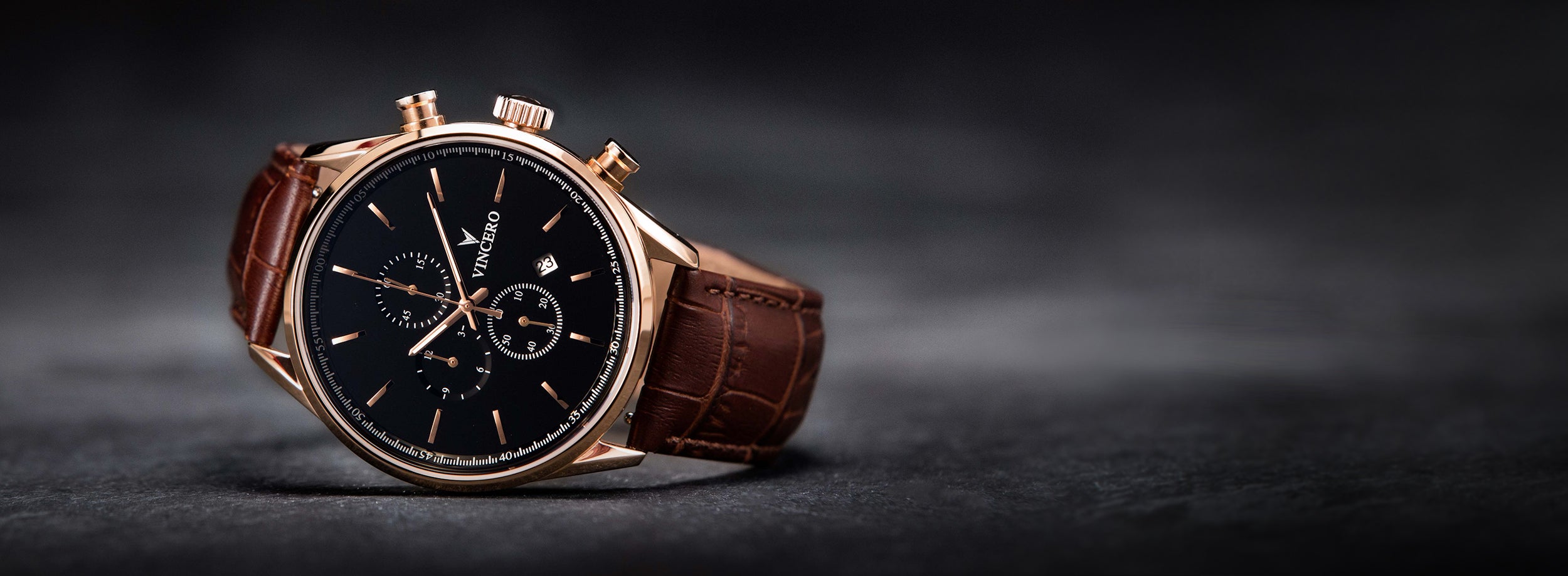 Close up of black and rose gold watch with brown leather strap in greyish black background