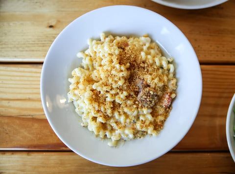 A classic macaroni and cheese is easily a comfort food for many, featuring a flatlay of a white plate of macaroni and cheese, garnished with breadcrumbs and powders, laid on top of a wooden table. Photo by Hayden Walker.