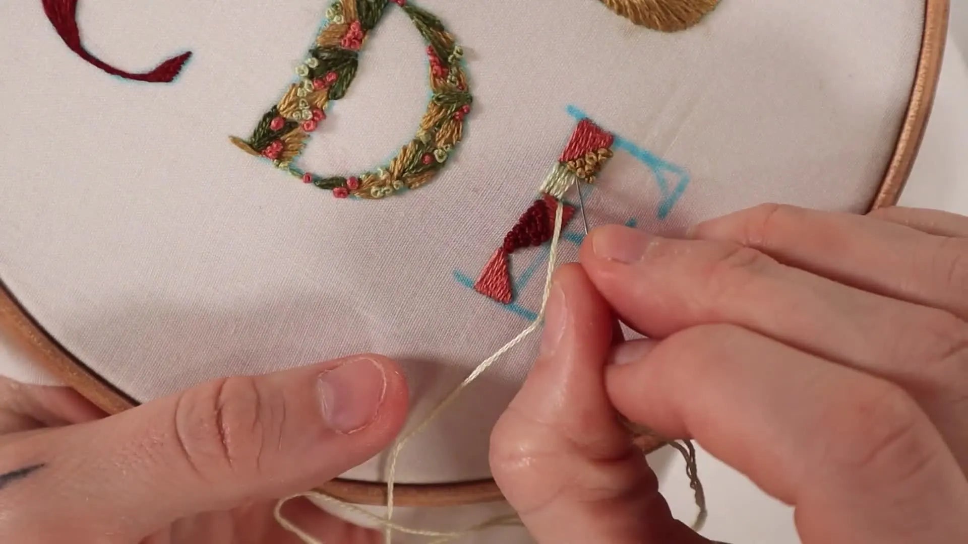 Hands filling geometric sections of letter 'E' with different colored satin stitches and French knots.