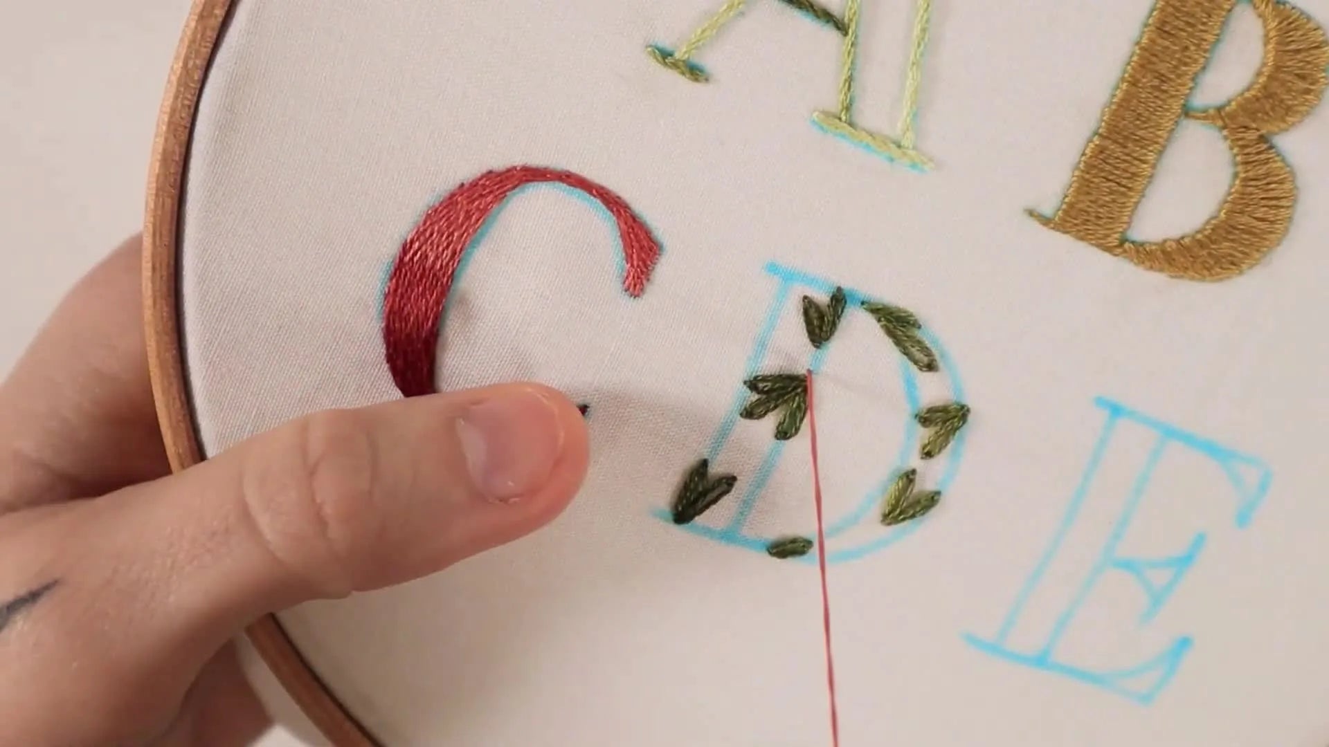 Hands adding pink French knots to the floral letter 'D' for flower buds.
