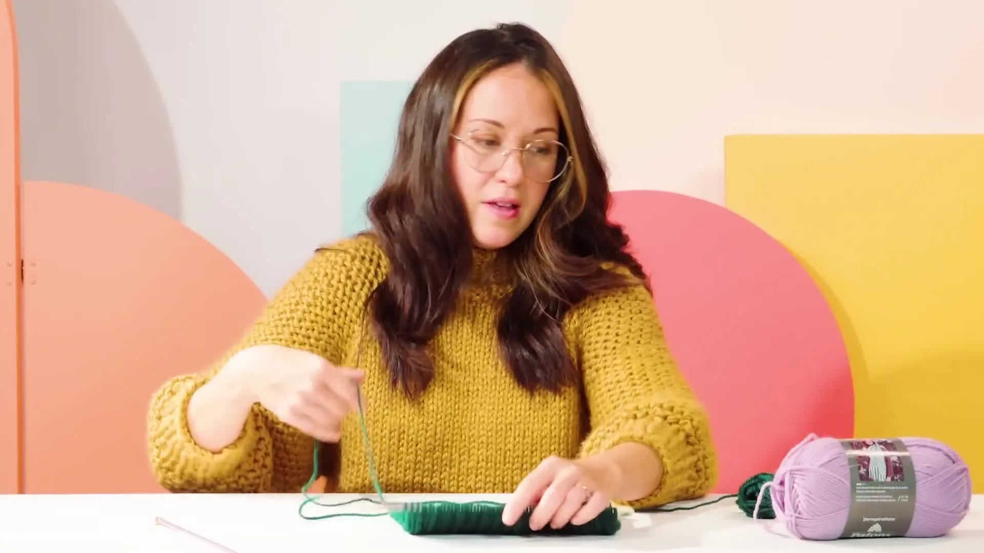 A completed section of green rib knit fabric on a knitting needle, with a new lilac yarn ready to be introduced.