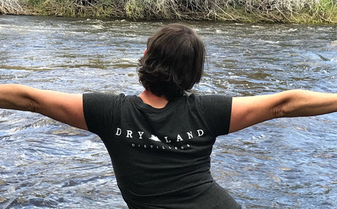 Woman with Dry Land Distillers t-shirt standing with arms up in the river