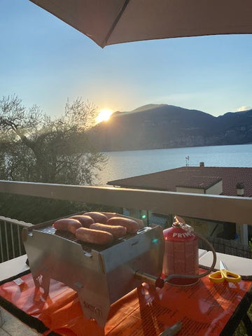 Faltbarer Gasgrill Skotti mit Würstchen auf dem Grillrost vor traumhafter Kulisse am Gardasee