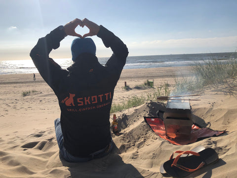 Mann grillt mit dem Skotti Gasgrill am Strand und formt ein Herz aus seinen Händen