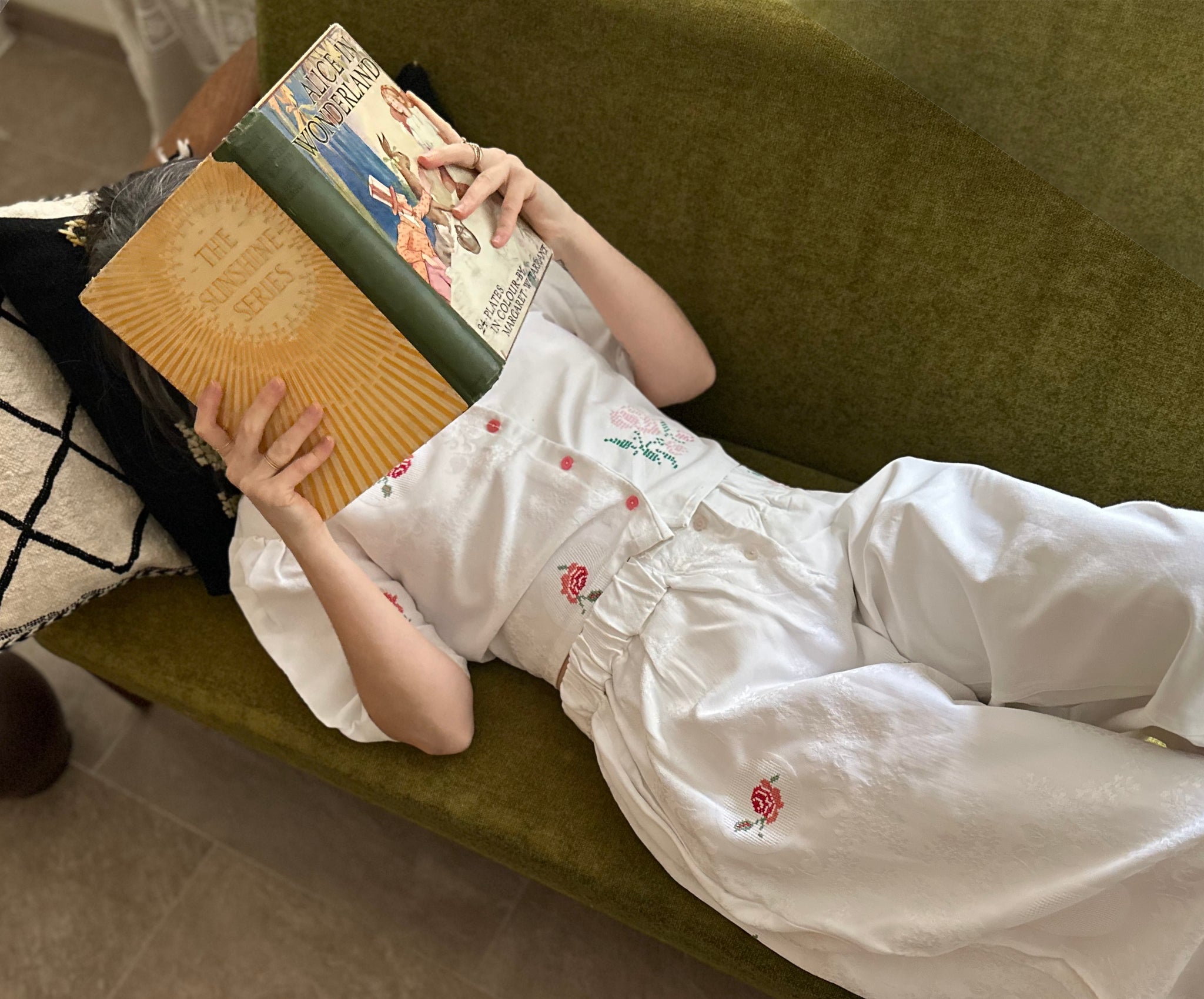 Vicki is lying on a green couch reading a large book, wearing a upcycled tablecloth co-ord set.