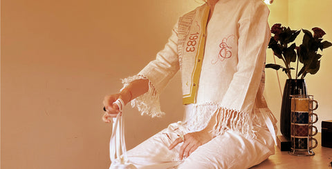 Vicki sits on a table wearing a 1983 tea towel shirt and trousers with a small bag in her hand. Behind is a vase of flowers