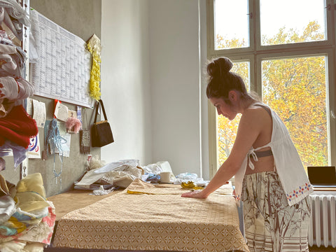 Vicki trägt ein Vintage-Kinderkleid als Oberteil und eine Hose aus Bettlaken, sie arbeitet an ihrem Schneidetisch in ihrem Atelier.