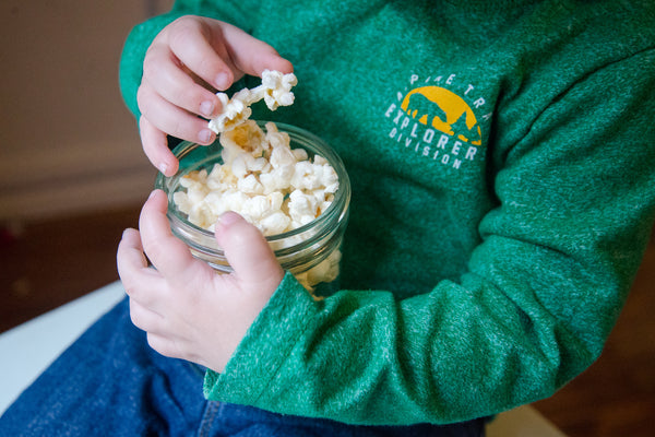 little boy wearing a green long sleeve shirt eating handfuls of fresh buttered popcorn from a mason jar