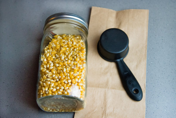 mason jar full of popcorn kernels next to a brown paper bag and a black measuring cup