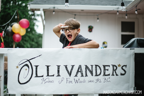 A young boy wearing Harry Potter glasses and pointing a wand and leaning over a hand painted sign for Ollivander's Wand Shop