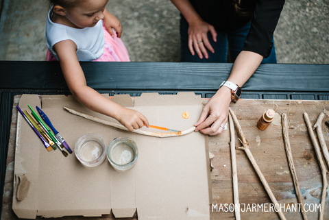 Young girl in a white and pink dress holding a paint brush and painting a hand carved wand with bright orange paint