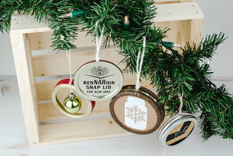 four DIY Christmas ornaments made out of used mason jar lids and rings hanging from an artificial evergreen bow in front of a natural wood crate