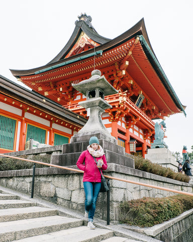 woman in pink jacket holding reusable mason jar tumbler and standing in front of a temple in Japan