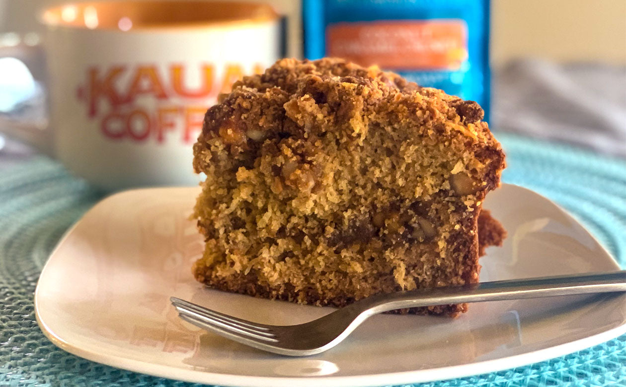 A slice of caramel crunch cake is in the foreground. A bag of kauai coffee coconut caramel crunch and a white mug are in the background 