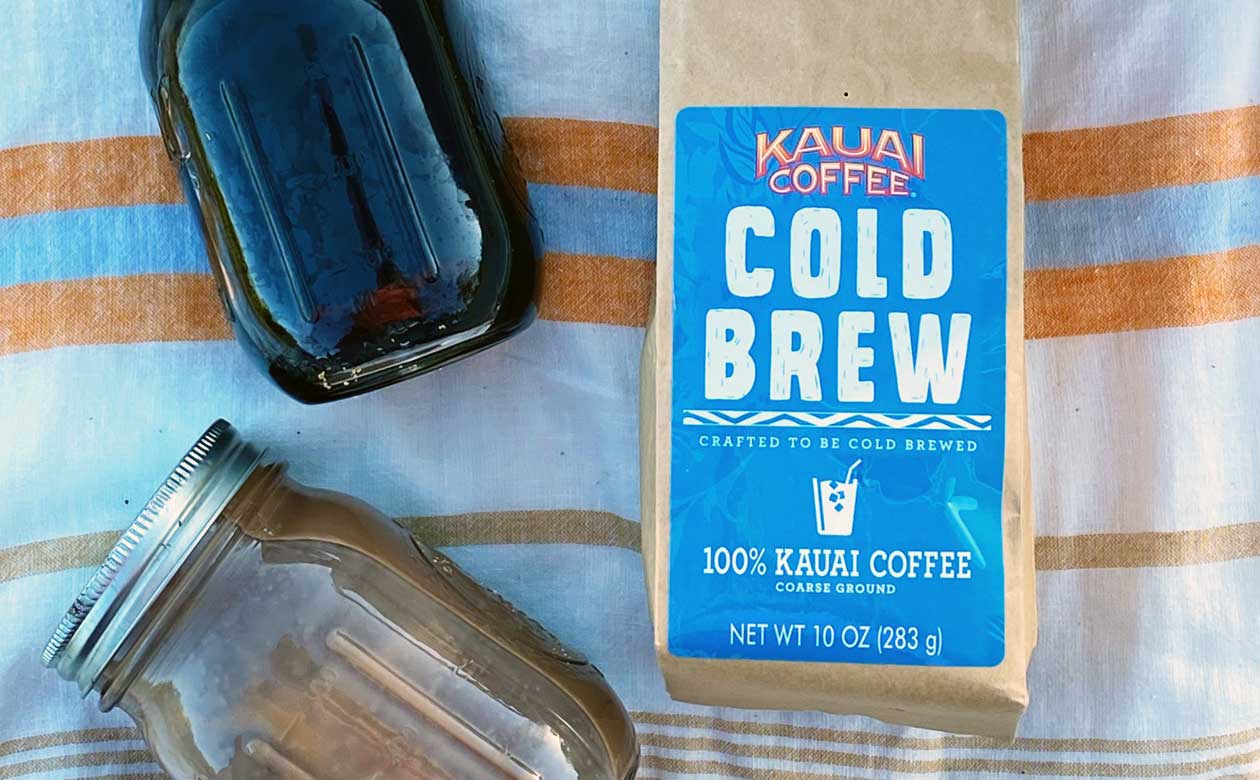 a bag of kauai coffee cold brew along with two mason jars full of coffee lay on a striped beach blanket