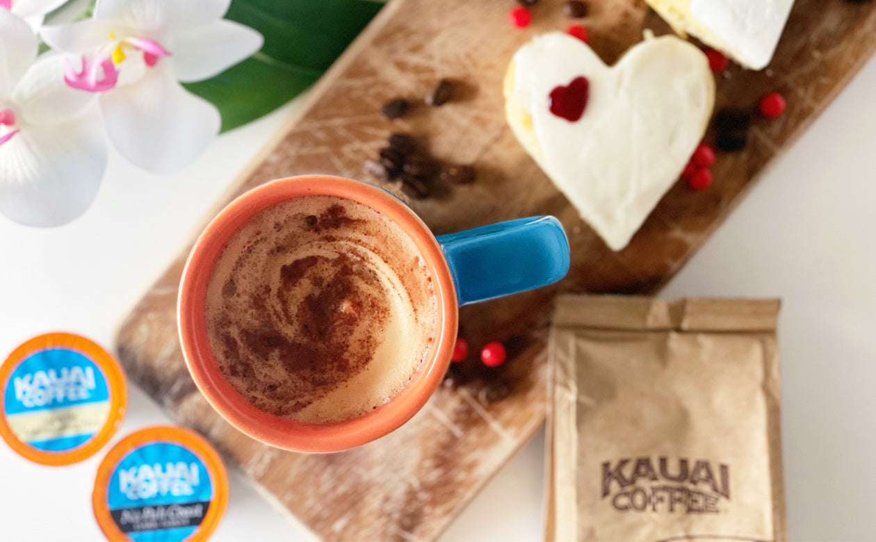 a mug of kauai coffee sits on a table with two scones. The scores are decorated with valentines' day hearts and frosting