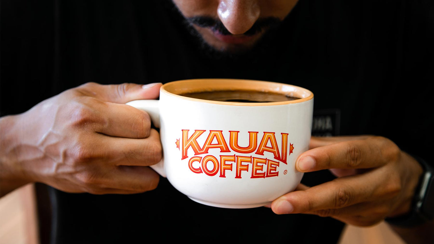 A close up image of a young man sipping black kauai coffee from a large, white mug that has the kauai coffee logo printed on it