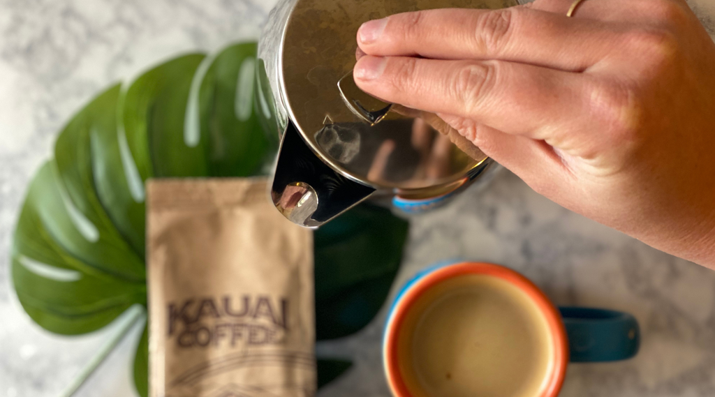 a hand presses the plunger on a silver french press brewer. A cup of coffee also sits on the white marble counter top