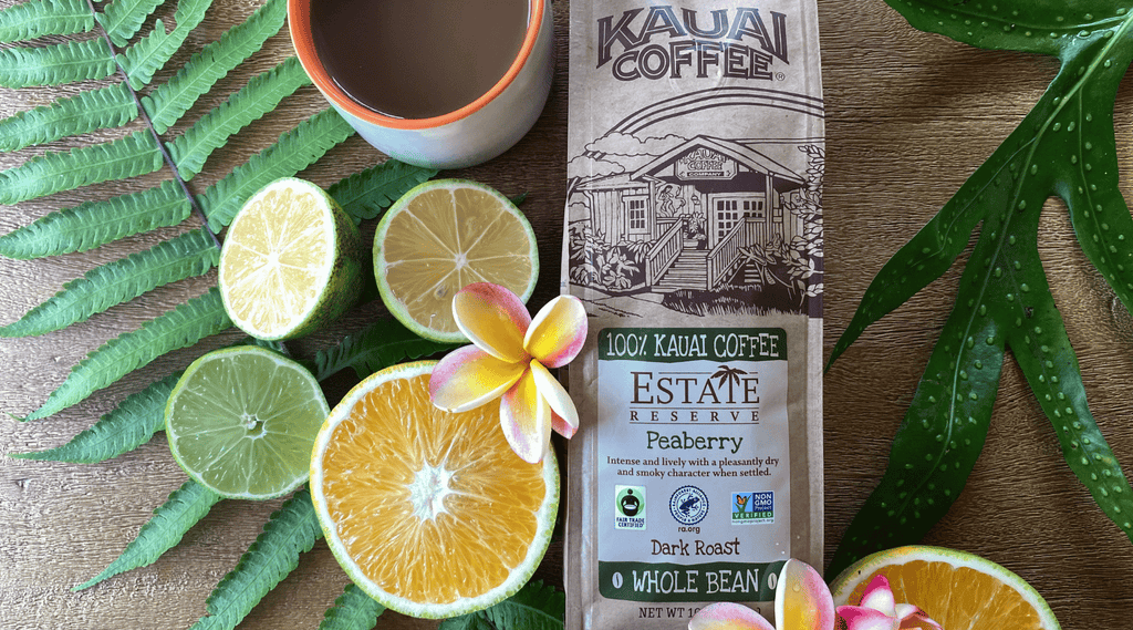 kauai coffee peaberry dark bag lays on a wooden surface with sliced citrus and tropical flora