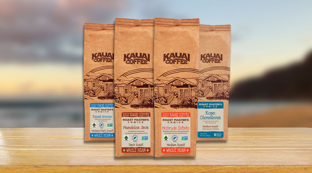 kauai coffee roast master's choice collection bags sitting on a wooden table. a blurred image of the na pali coast is visible in the background