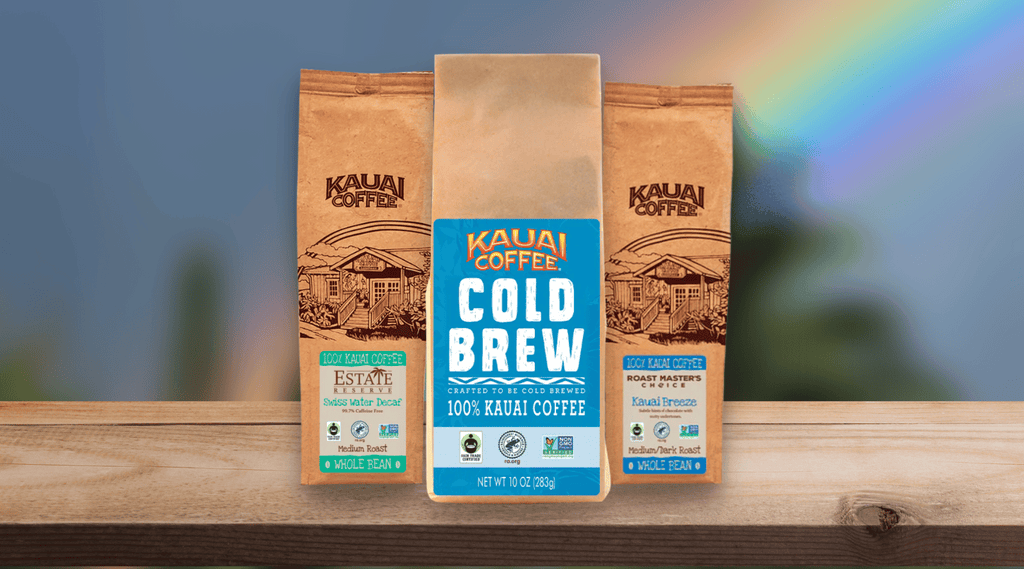 100% Kauai Coffee best for cold brewing. A bag of cold brew, estate reserve decaf and kauai breeze are shown on a table. A rainbow is visible in the background