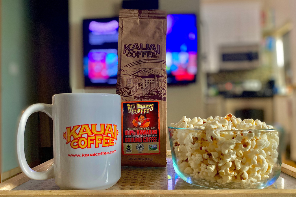 kauai coffee big braddah sits on a tv tray in front of a big screen tv ready to play a movie. There is a bowl of popcorn and a fresh cup of coffee on the table next to the bag of coffee