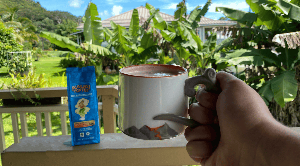 a hand holds a white mug filled with a cafe latte. A tropical scene and a bag of kauai coffee brewberry crumble is visible in the background