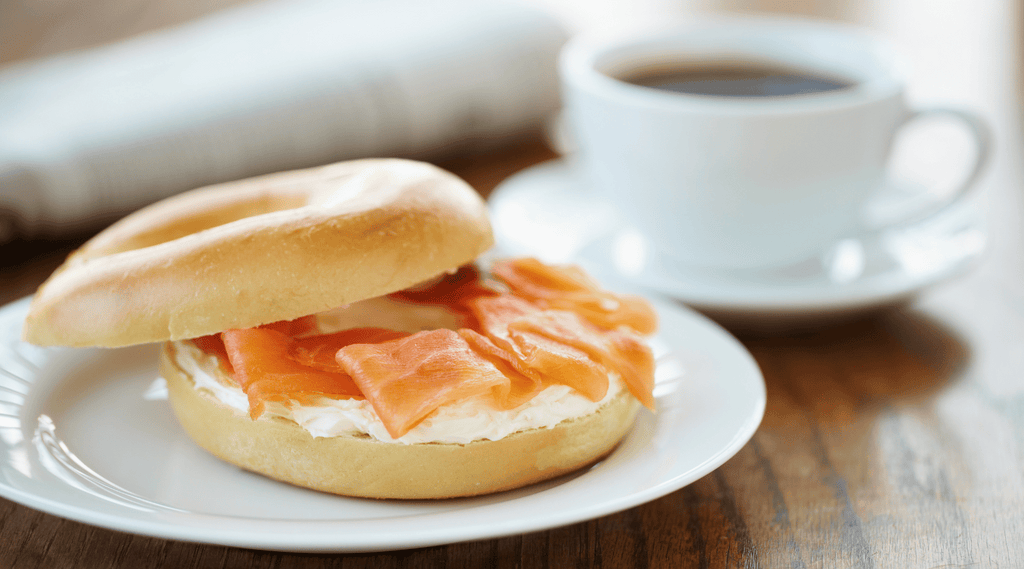 a sliced plain bagel topped with cream cheese and smoked salmon sits on a white plate. A white mug full of coffee sits in the background. 