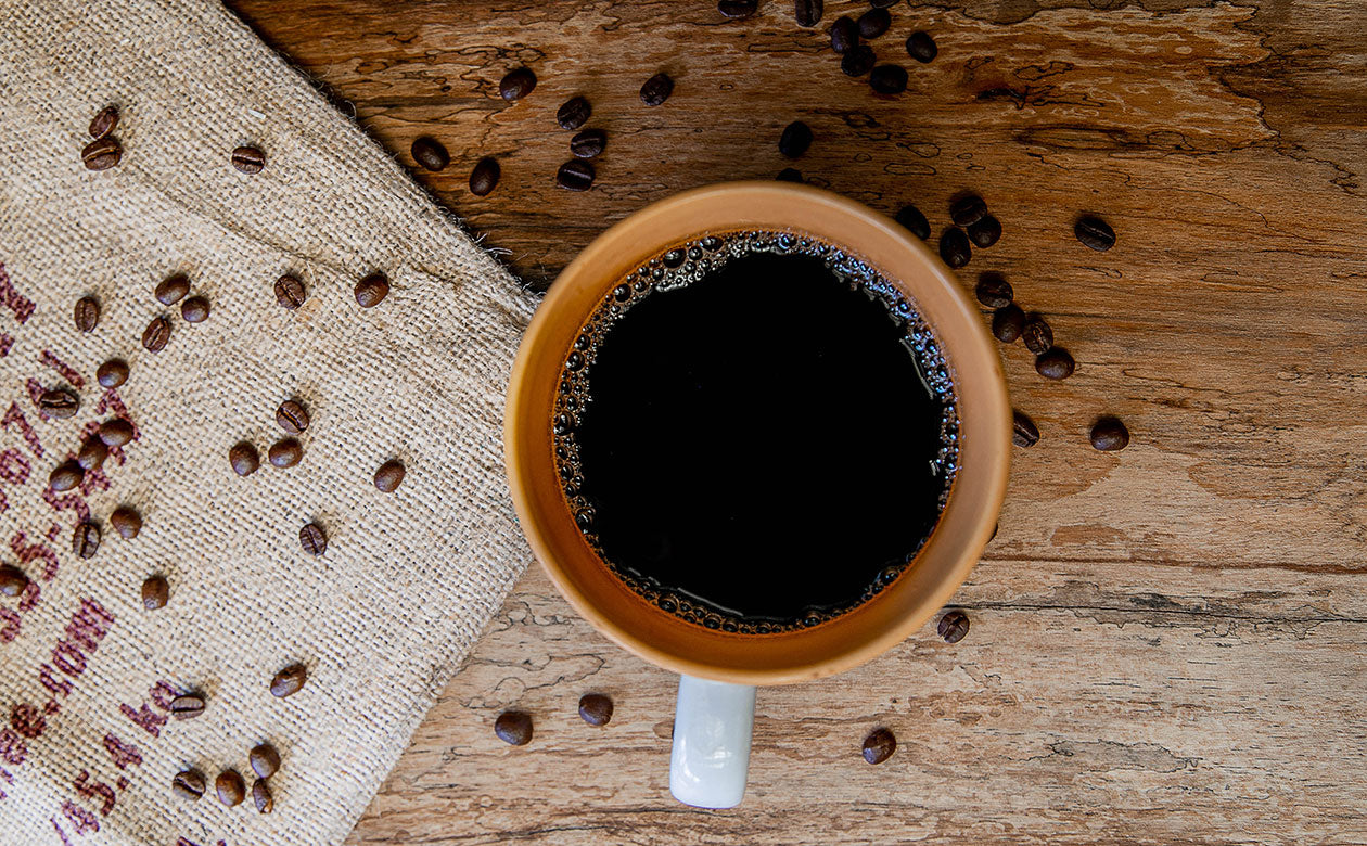 a birds-eye view of a large white coffee mug sitting on a burlap coffee bag. Some coffee beans are scattered across the table