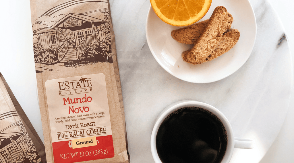 a bag of kauai coffee estate reserve mundo novo sits on a white granite table with a sweet breakfast spread of biscotti and brewed coffee